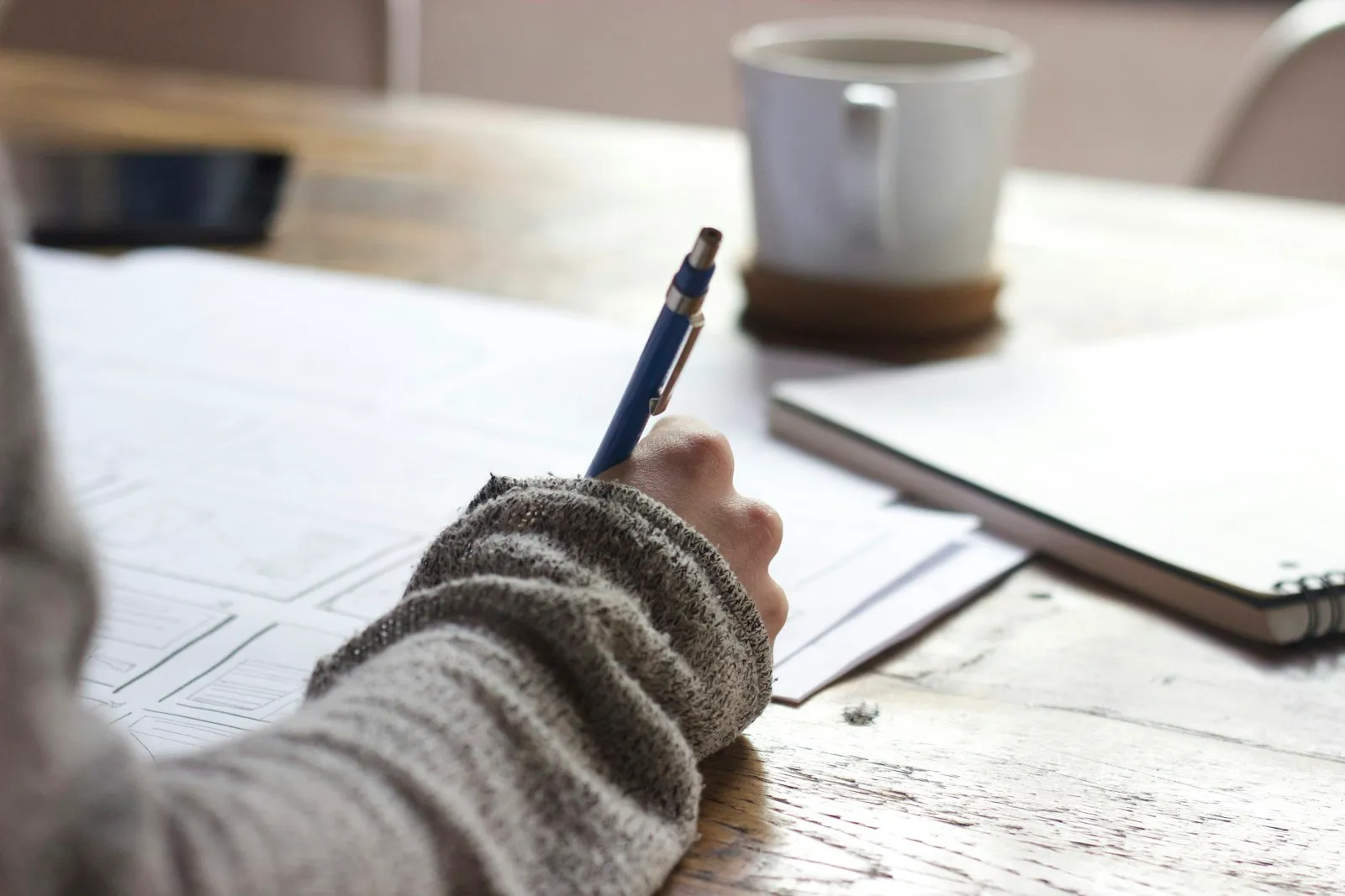 Person writing on a table beside a ceramic mug